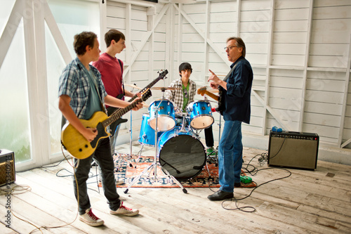 Mid adult man talking to three teenage boys playing with musical instruments inside a garage. photo