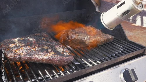 Flame grilling beef on a barbecue grill in slow motion. Fire shoots out of a flame thrower over sizzling smoked meats. Action shot would be perfect for videos on summertime grilling competitions.