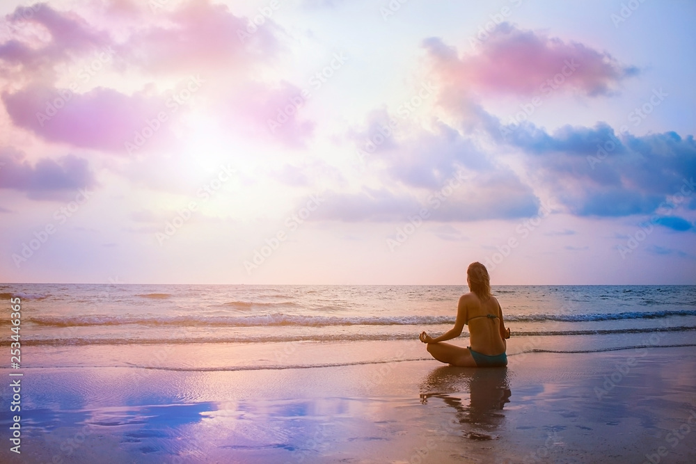 Young beautiful woman sitting on sunset sea beach and practicing zen yoga in lotus pose. Evening meditation with view on surrealistic sky and ocean. Reflection on wet sand