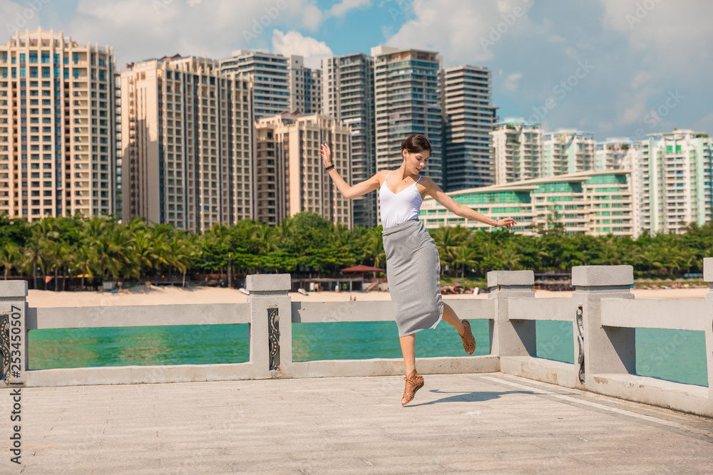 Cheerful and beautiful girl having fun on vacation, jumping on the pier on background of city. In China, Hainan city of Sanya. Town Sanya is popular tourist destination in China. View of island in bay