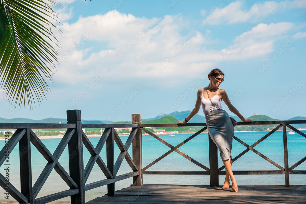 Cheerful and beautiful girl resting fun in vacation, on the pier on background of city. In China, Hainan city of Sanya. View of island in bay. Town Sanya is popular tourist destination in China Cheerful and beautiful girl resting fun in vacation, on the pier on background of city. In China, Hainan city of Sanya. View of island in bay. Town Sanya is popular tourist destination in China