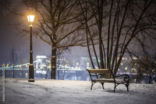 Budapest, Hungary - Bench and lamp post in a snowy park at Buda district with Szechenyi Chain Bridge at background during heavy snowing at winter time