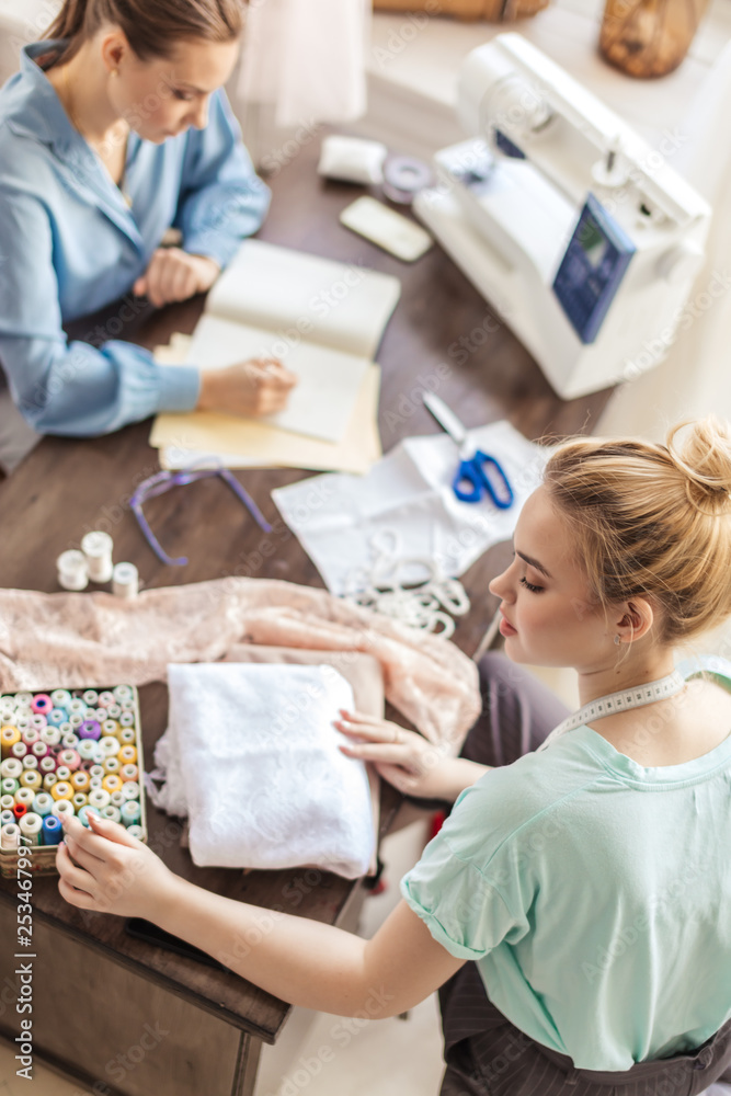 Obraz premium 2 young beautiful female dressmakers cooperating in their own studio working as a team on new order. Small Business, partnership and friendship concept