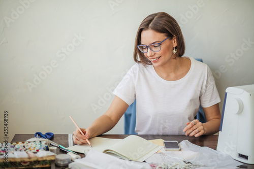 Skilled beautiful dressmaker in fashion atelier in stylish eyeglasses and white casual t-shirt workingstanding at desktop and drawing sketch for collection of textile clothing - 