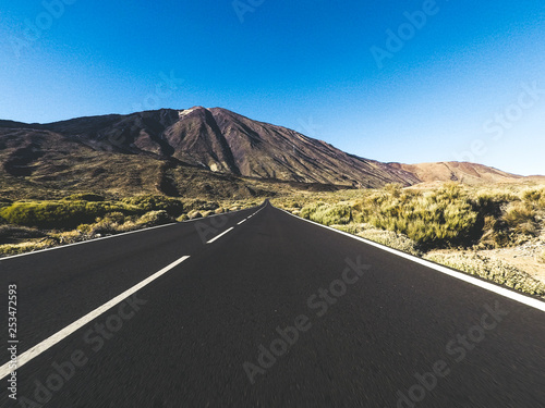 Long way road at the mountain with vulcan mount in front and blue clear sky - ground point of view with black asphalt and white lines - driving and travel concept