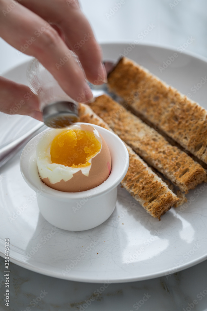 Fototapeta premium Frau salzt und isst gekochtes Ei im Eierbecher mit Eierlöffel, Salzstreuer und Toast Brot beim Frühstück