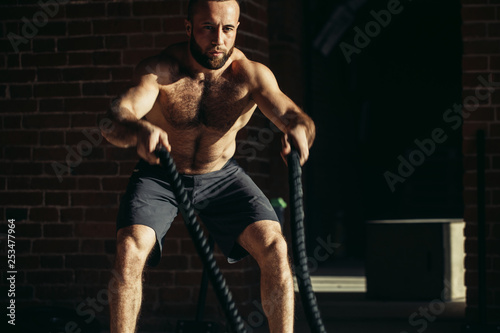 Athletic young man with battle rope doing exercise in functional training fitness gym. Rope helps engage all muscle groups at the same time, while ensuring freedom of movement.