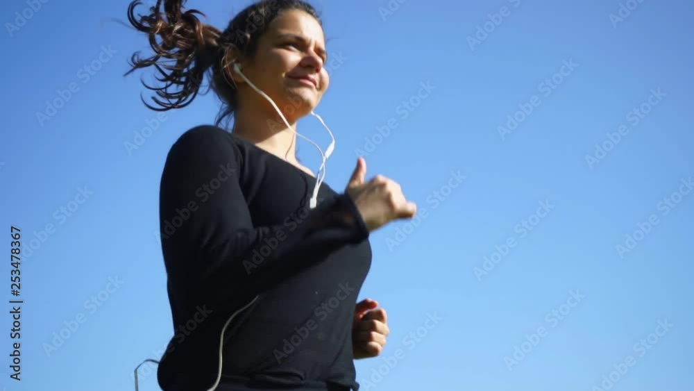 Sporty girl running against blue sky. Low angle view of athletic young woman in earphones jogging at sunny day, slow motion. Workout concept Sporty girl running against blue sky. Low angle view of athletic young woman in earphones jogging at sunny day, slow motion. Workout concept