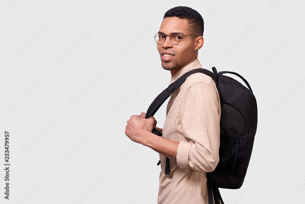 Indoors portrait of handsome young Afro man student with backpack standing over whiet studio background. Young businessman wearing casual beige shirt and round eyewear. People concept Indoors portrait of handsome young Afro man student with backpack standing over whiet studio background. Young businessman wearing casual beige shirt and round eyewear. People concept