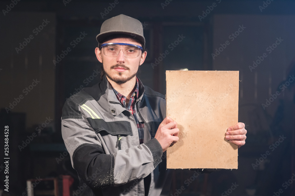 Portrait of a young carpenter in overalls and goggles with a mock up board holding a blank sign in his hands in his home workshop Portrait of a young carpenter in overalls and goggles with a mock up board holding a blank sign in his hands in his home workshop