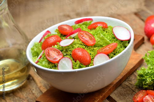 Close-up of green salad Lollo Biondo with tomatoes and radishes in a white bowl on wooden table, a pitcher of oil in the background