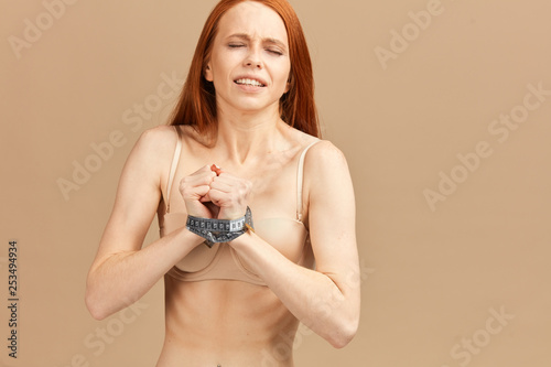 Unhappy woman with ginger hair, suffering from eating disorders posing isolated over beige background with hands tied with measuring tape, wants to get free from mental prison