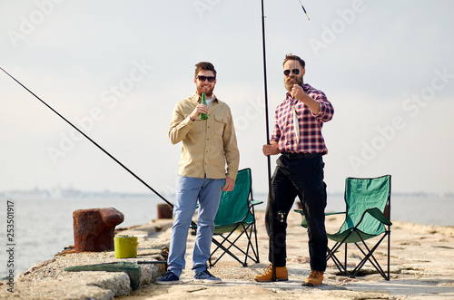 leisure and people concept - happy friends with fishing rods, fish and beer on pier at sea