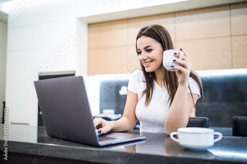 Portrait of a beautiful young woman with coffee cup using laptop in the kitchen at home