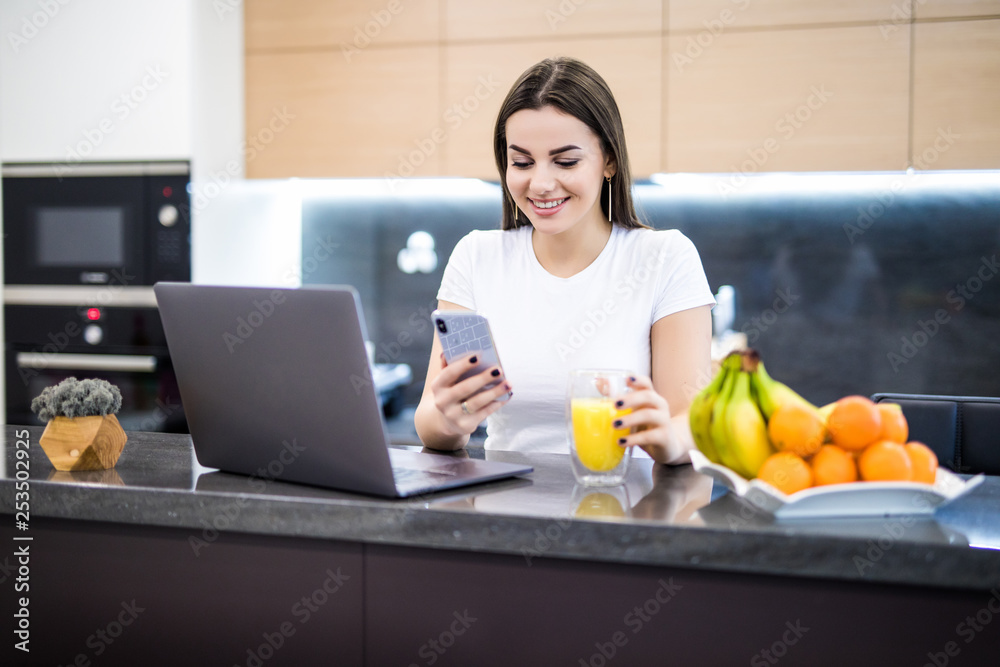 Young woman student work on computer while talking on smart phone, use laptop sitting at table in kitchen, freelancer working on notebook Young woman student work on computer while talking on smart phone, use laptop sitting at table in kitchen, freelancer working on notebook