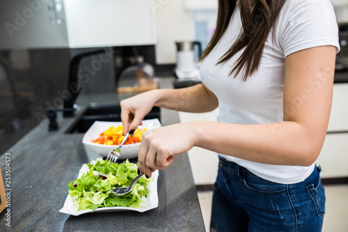 Beautiful young girl in a white shirt prepares healthy food with greens. Woman mixed salad in kitchen.