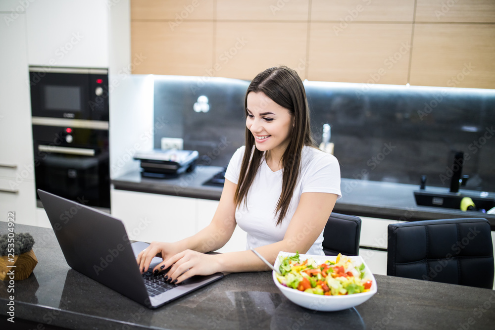 Cheerful young woman eating salad from a bowl while standing on a kitchen and watching movie on laptop