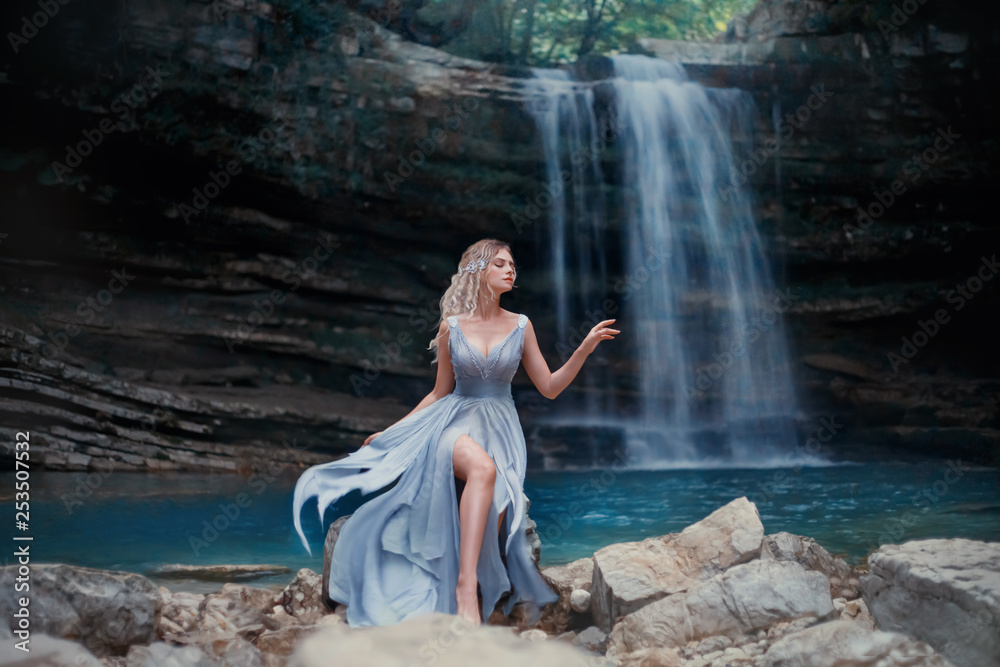 A curly blonde girl in a luxurious blue dress sits on white stones against the backdrop of a fabulous landscape. River Mermaid near the lake with a waterfall. Art Photography