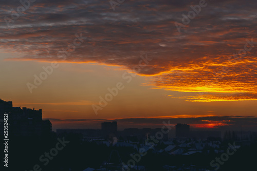 Cityscape with vivid fiery dawn. Amazing warm dramatic cloudy sky above dark silhouettes of city building roofs. Orange sunlight. Atmospheric background of sunrise in overcast weather. Copy space.