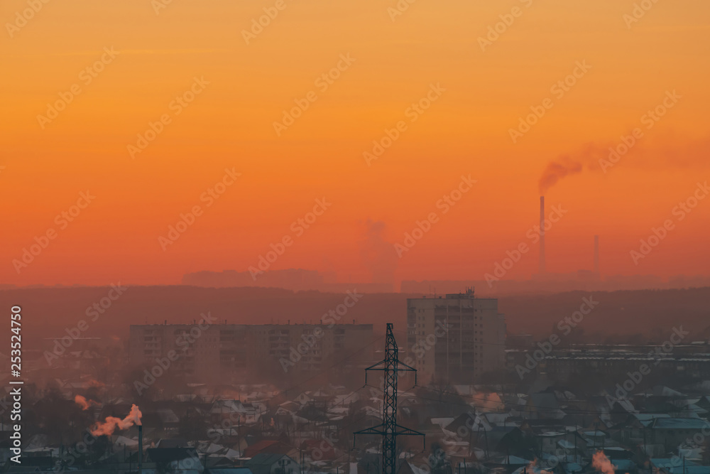 Power lines in city on dawn. Silhouettes of urban buildings among smog on sunrise. Cables of high voltage on warm orange yellow sky. Power industry at sunset. City power supply. Mist urban background.