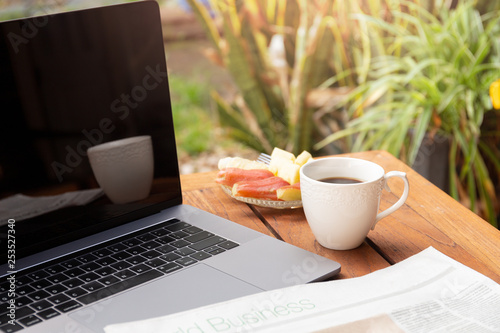 Coffee mug and news paper with fresh fruit and laptop on wooden table.