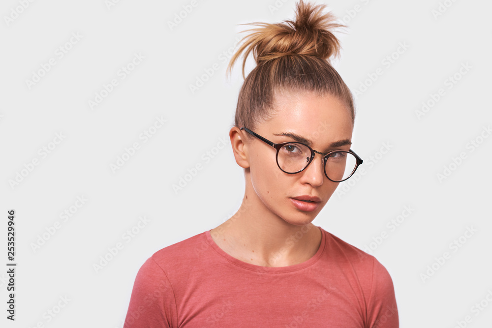 Attractive blonde young woman wears spectacles and casual pink blouse, looks seriously directly into camera, poses against white studio background. People emotions concept
