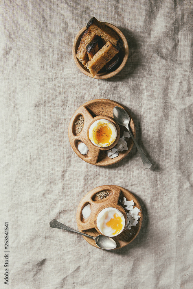 Breakfast with soft boiled eggs, served in wooden egg cup with salt, pepper and toasted bread in row over linen tablecloth. Flat lay, space