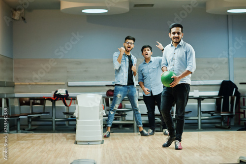 South asian man in jeans shirt standing at bowling alley with ball on hands. Guy is preparing for a throw. Friends support him loudly.