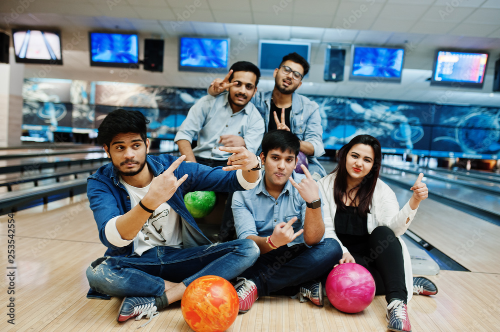 Group of five south asian peoples having rest and fun at bowling club, sitting on bowling alley with balls on hands.