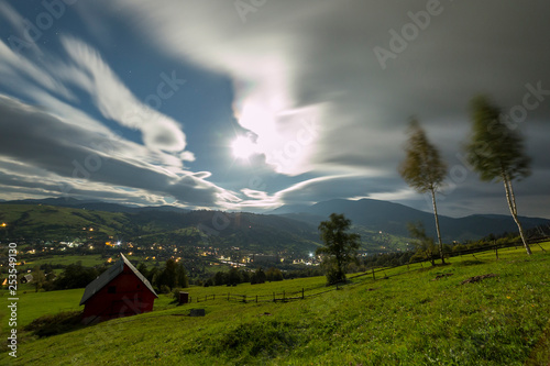 Summer night mountain panorama. Small house cottage on steep mountain slope on dramatic evening sky background, bright road with moving cars and resort dwelling lights in valley.