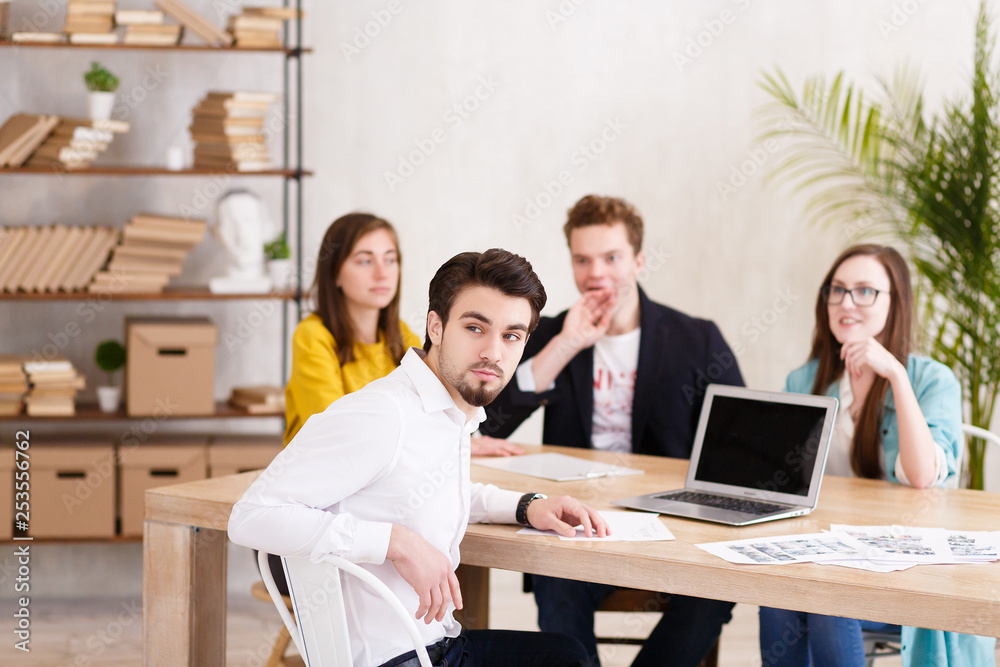 Fototapeta premium Portrait of handsome successful businessman in white shirt sitting at conference table against the background of blurry employees of his company