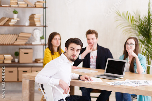 Portrait of handsome successful businessman in white shirt sitting at conference table against the background of blurry employees of his company