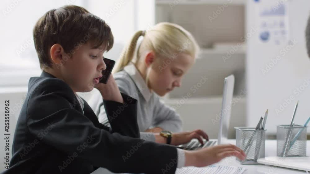 Panning of Caucasian boy and girl sitting at desk in office. Girl typing on laptop keyboard, boy talking on phone Panning of Caucasian boy and girl sitting at desk in office. Girl typing on laptop keyboard, boy talking on phone