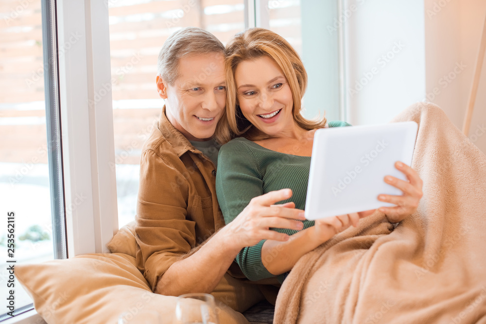 smiling couple using digital tablet together while sitting by window