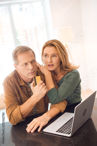 thoughtful couple standing by laptop while husband holding credit card