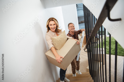 Φωτογραφία smiling happy couple carrying things upstairs and looking at camera