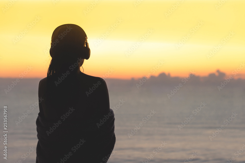 Silhouette of naked woman with wet hair wrapped in a blanket after swimming. Female in the headphones standing on the beach opposite sea.