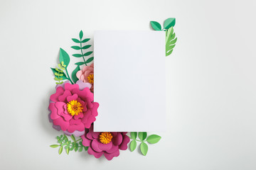 top view of white blank card near pink paper flowers and green leaves on grey background