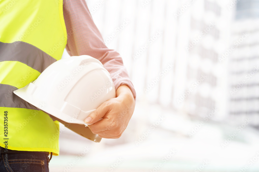 Naklejka premium Close up front view of engineering male construction worker holding safety white helmet and wear reflective clothing for the safety of the work operation. outdoor of building background.