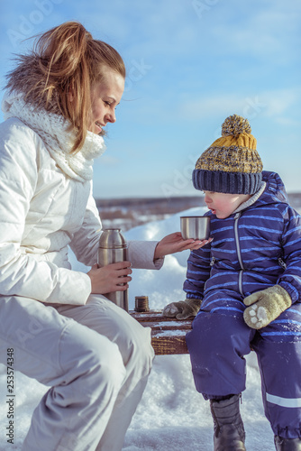 A young mother gives a child a boy 3-5 years old to drink a cup of hot tea or soup, a drink to warm on a cold winter day outside. Camping on a winter day off.