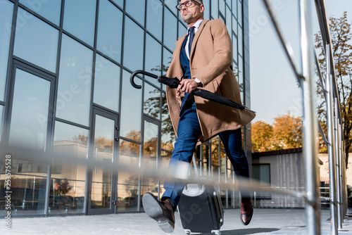 handsome businessman in glasses and beige coat running with suitcase and umbrella on street