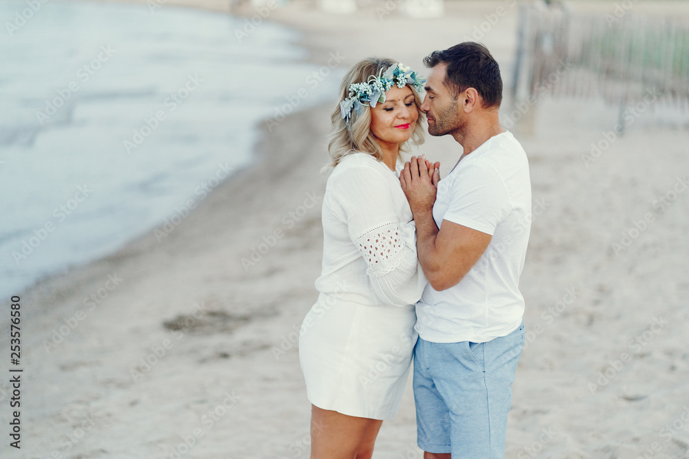 elegant and stylish woman with light curly hair dressed in a white blouse walking on the beach near water along with her elegant man in a white t-shirt and in a  blue shorts
