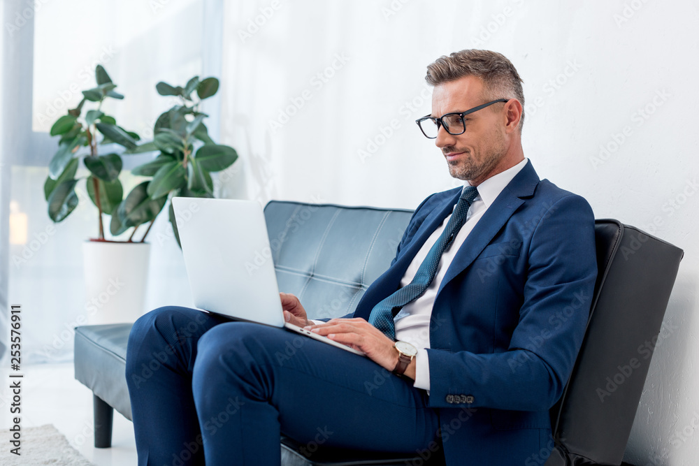 cheerful businessman in suit using laptop while sitting on sofa