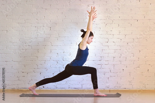 The girl is a professional instructor of hatha yoga practicing asanas in the room against the background of a white brick wall. Virabhadrasana 1 (Warrior Pose 1).