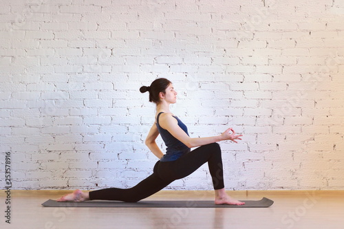 The girl is a professional instructor of hatha yoga practicing asanas in the room against the background of a white brick wall. Ashwa Sanchalanasana variation (Rider Pose).