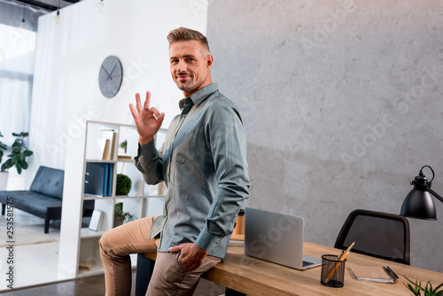 Εκτύπωση καμβά happy businessman showing ok sign while sitting with hand in pocket in office
