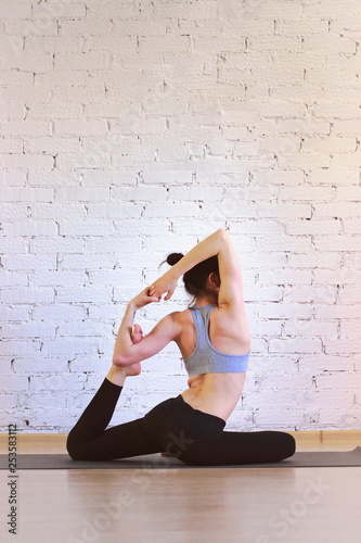 The girl is a professional instructor of hatha yoga practicing asanas in the room against the background of a white brick wall. Eka Pada Rajakapotasana (King Pigeon Pose).