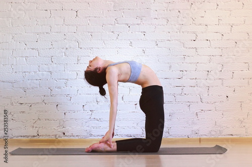 The girl is a professional instructor of hatha yoga practicing asanas in the room against the background of a white brick wall. Ustrasana (Camel Pose).