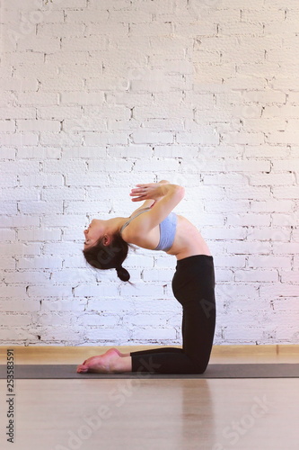 The girl is a professional instructor of hatha yoga practicing asanas in the room against the background of a white brick wall. Ustrasana (Camel Pose).