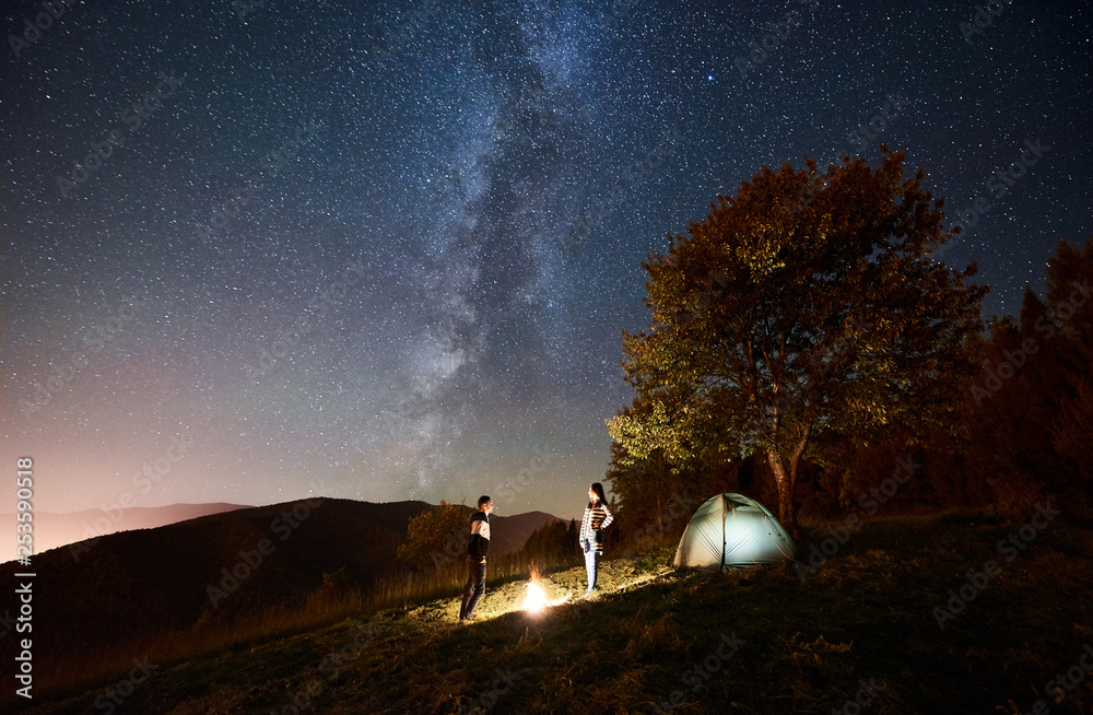 Young happy couple tourists standing at bonfire near illuminated tent under amazing night sky full of stars and Milky way. On the background beautiful starry sky, mountains and big tree